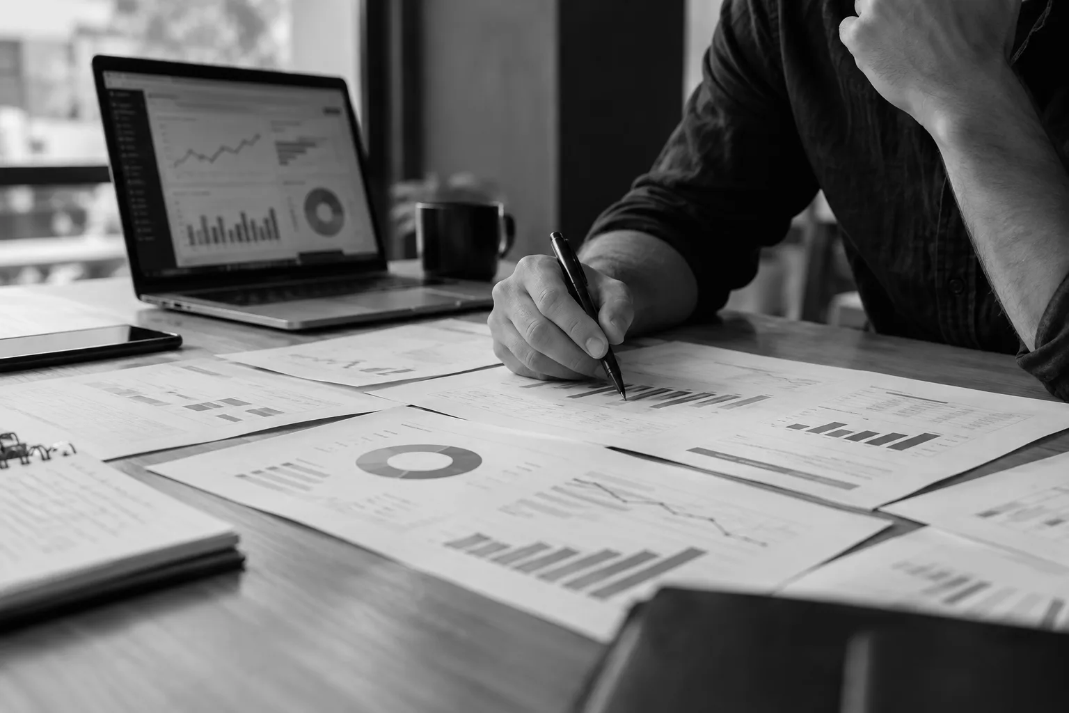 An owner at a desk marking up printed marketing reports, with a performance dashboard open on a laptop
